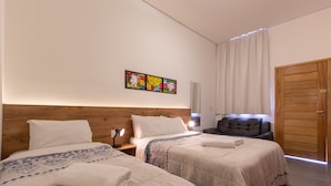Ein Hotelzimmer mit zwei Betten, einem Holzkopfstück, einem Nachttisch mit Lampe, einem schwarzen Ledersessel, einer Holztür und einem farbenfrohen Kunstwerk an der Wand.