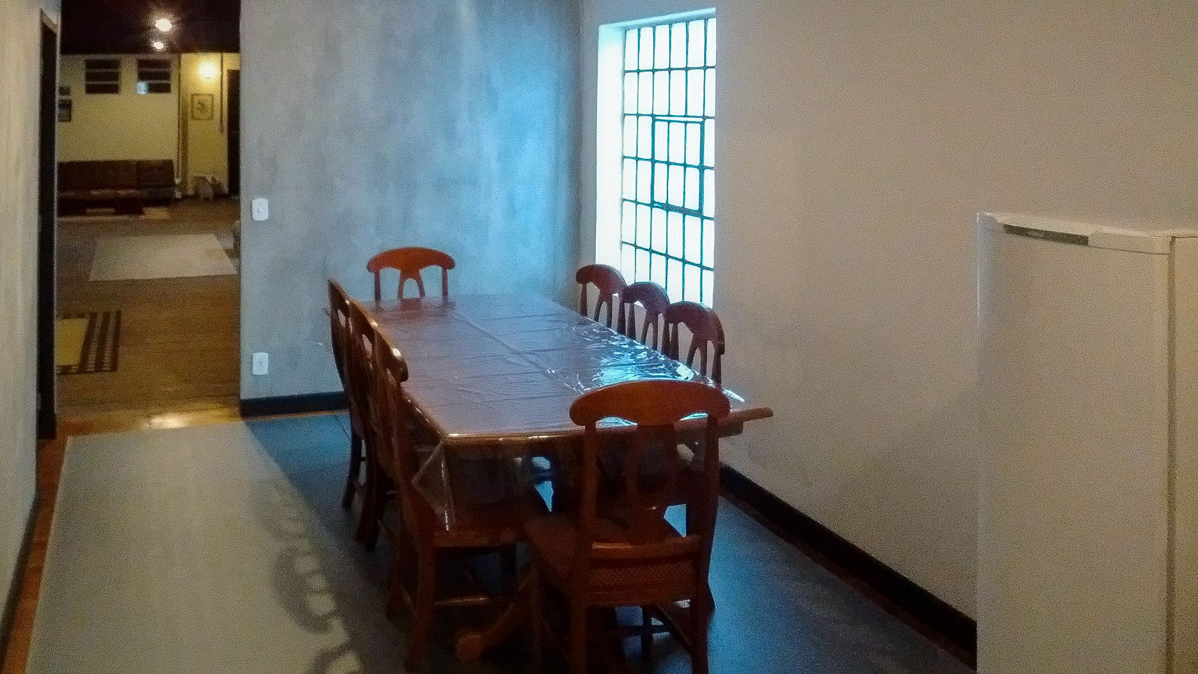 A dining area with a long table and chairs, a window with bars, and a door leading to another room.