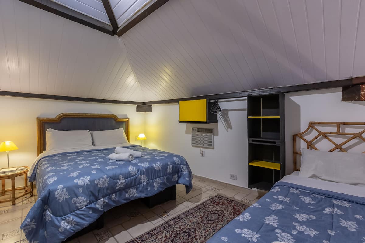 A bedroom with two beds, a wooden headboard, a small bedside table with a lamp, a wall-mounted TV, and a sliding shelf.