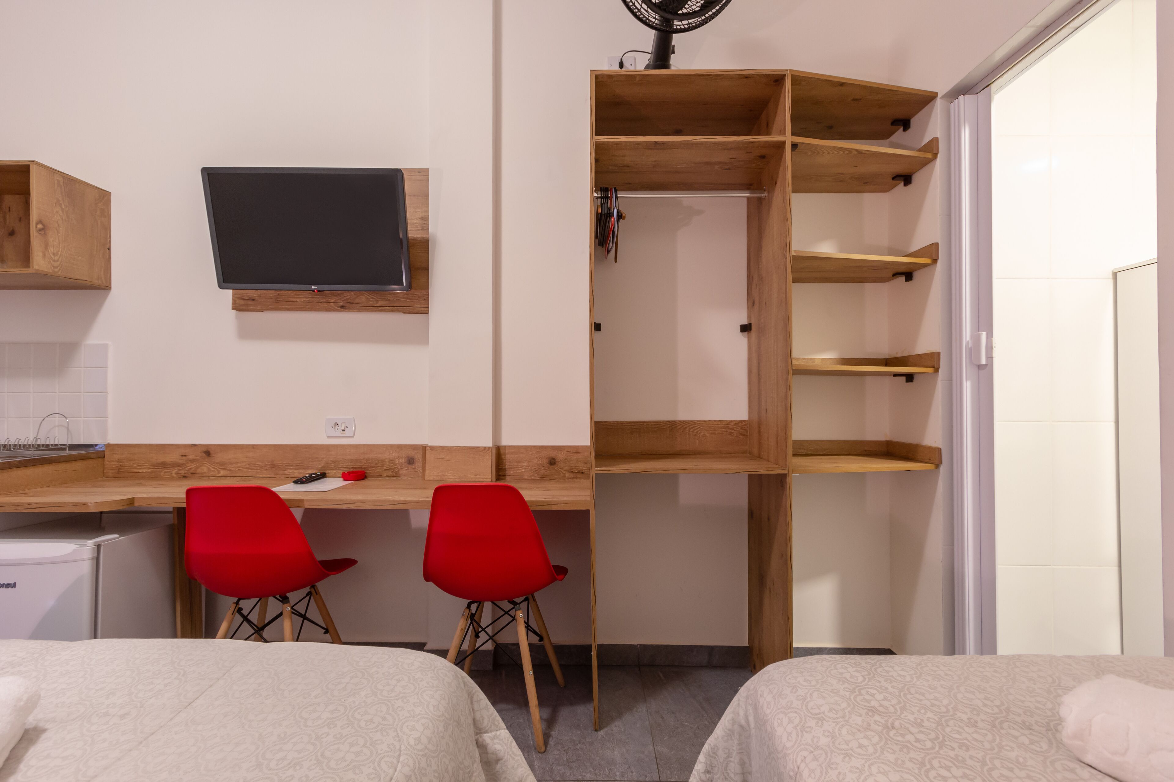 A bedroom with two beds, a desk with two red chairs, a wooden shelving unit, and a television on the wall.