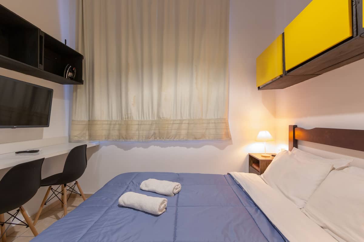 A hotel room with a bed, a desk with a chair, a TV, a window with curtains, a nightstand with a lamp, and a yellow cabinet.