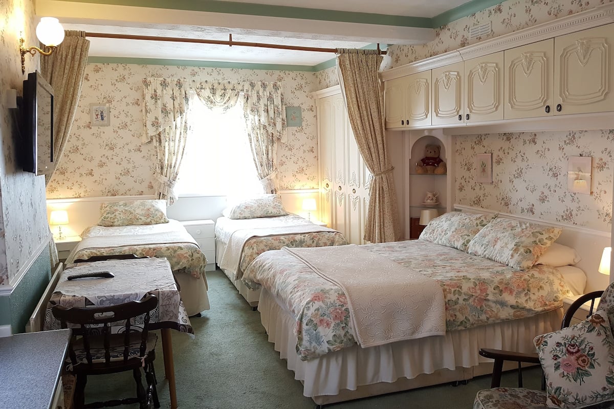 A room with two beds, a desk, and a chair. There is a window with curtains, a wall-mounted lamp, and a teddy bear on a shelf.