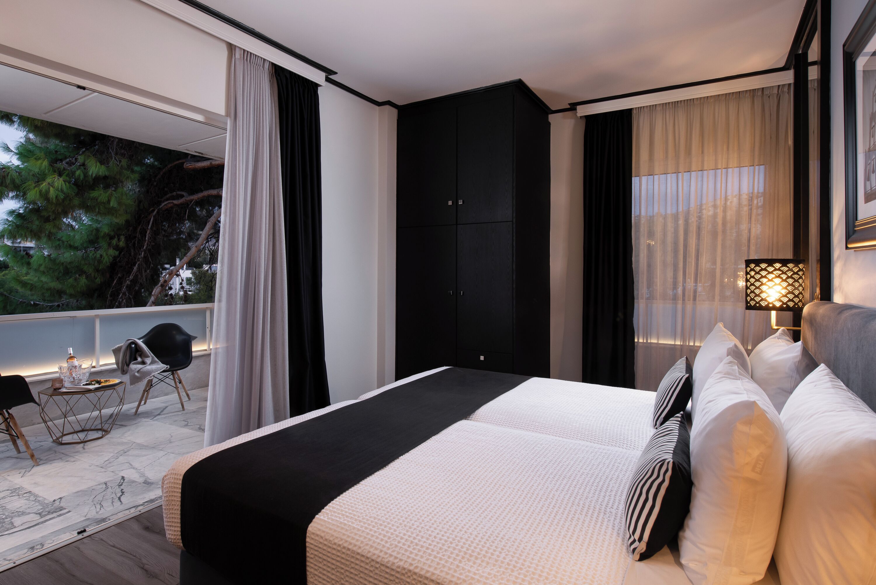 Une chambre d’hôtel moderne dotée d’un grand lit, d’un balcon offrant une vue sur les arbres et d’un coin repas avec une table et des chaises.