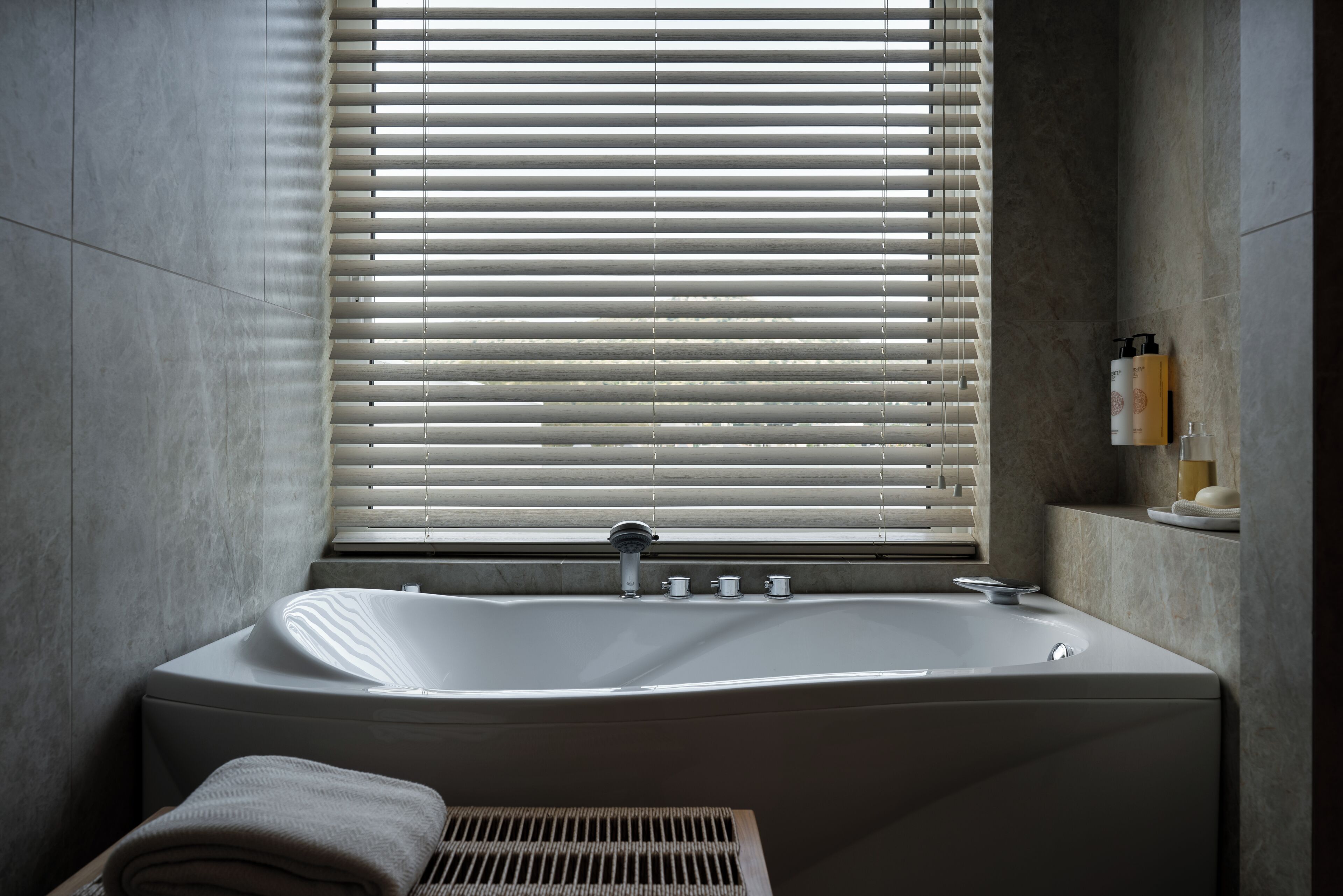 A modern bathroom with a bathtub, a window with blinds, and a towel on a bench.