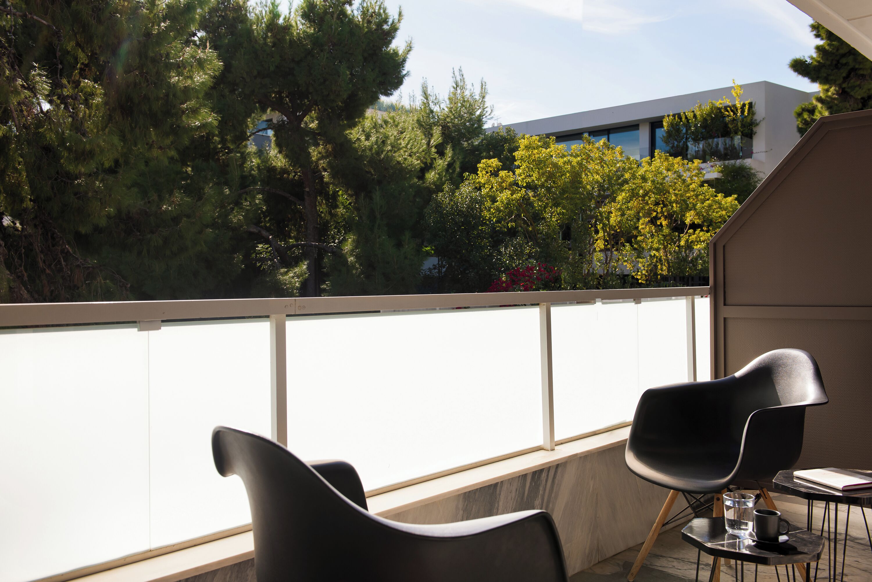 Un balcon avec deux chaises noires et une petite table, d’où l’on peut admirer un immeuble et des arbres.