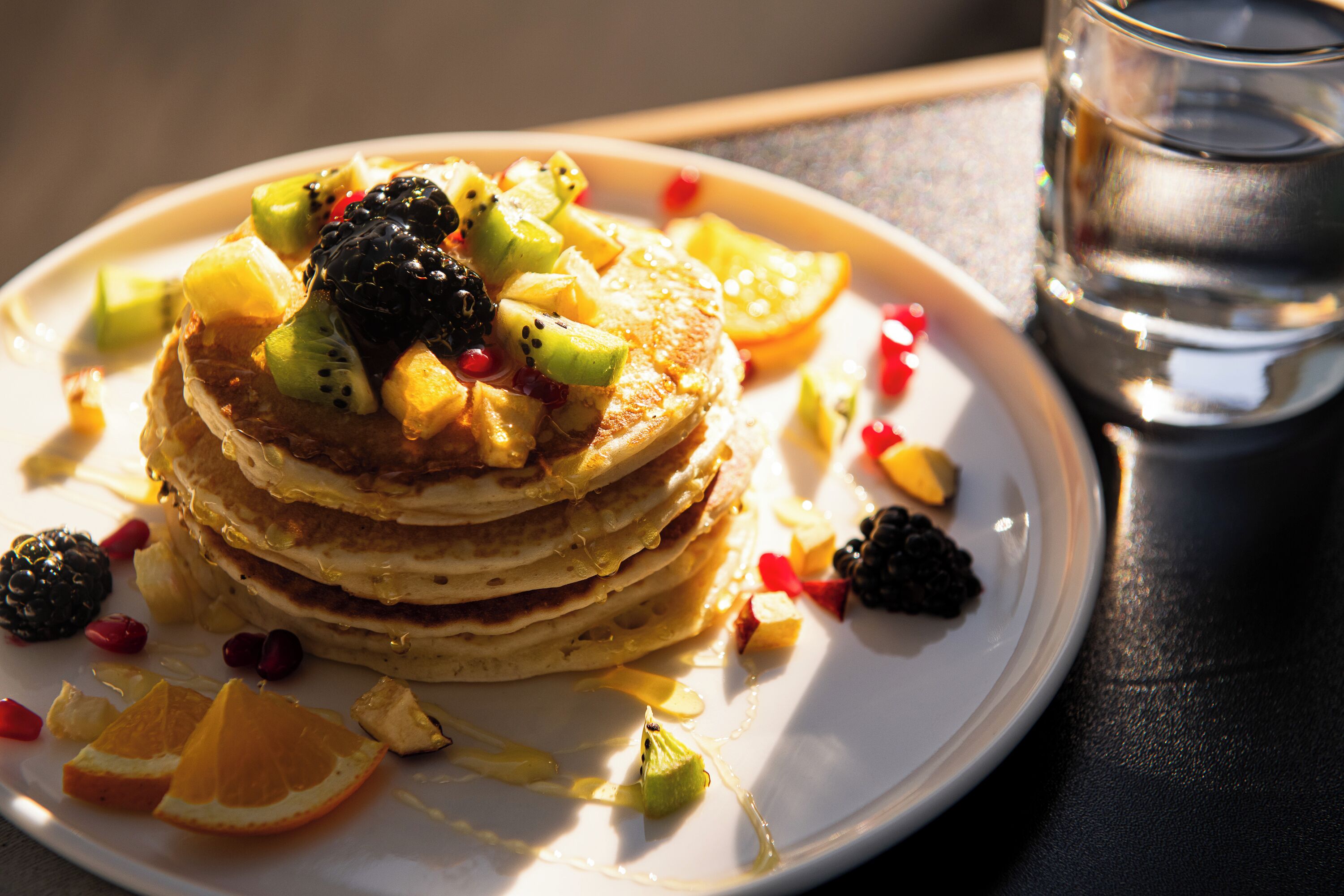 A stack of pancakes topped with fruit and syrup, served on a plate with a glass of water beside it.