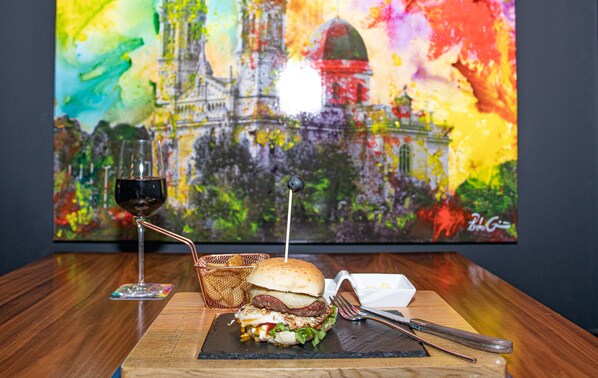 Una comida con una hamburguesa, una copa de vino tinto y un cuadro colorido de fondo.