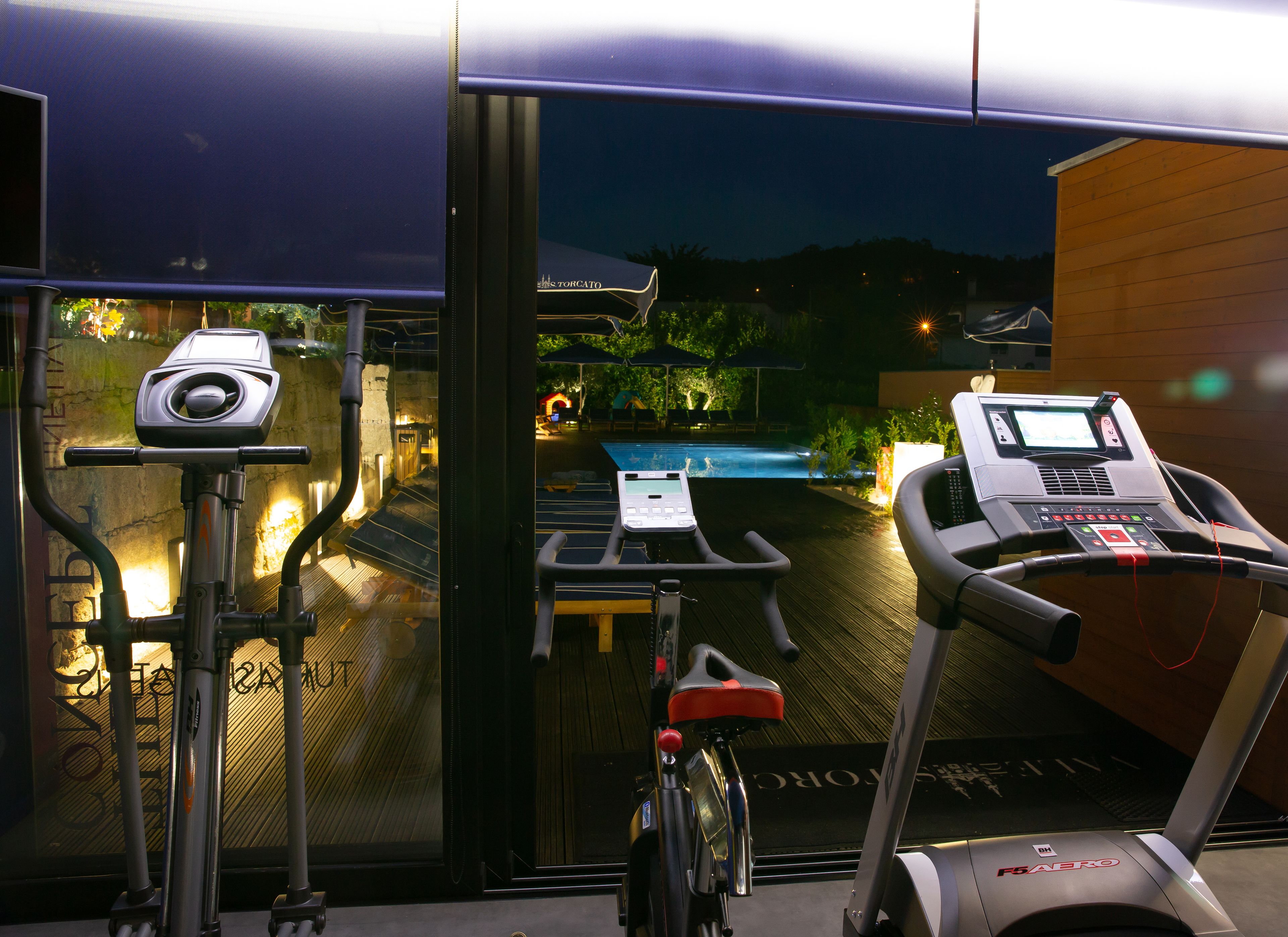 Une salle de sport intérieure équipée de matériel d’exercice, avec une piscine et une vue sur l’extérieur, de nuit.