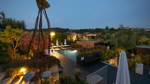 Zona de piscina en la azotea con sillones de descanso y sombrillas, rodeada de vegetación exuberante y vistas a una colina.