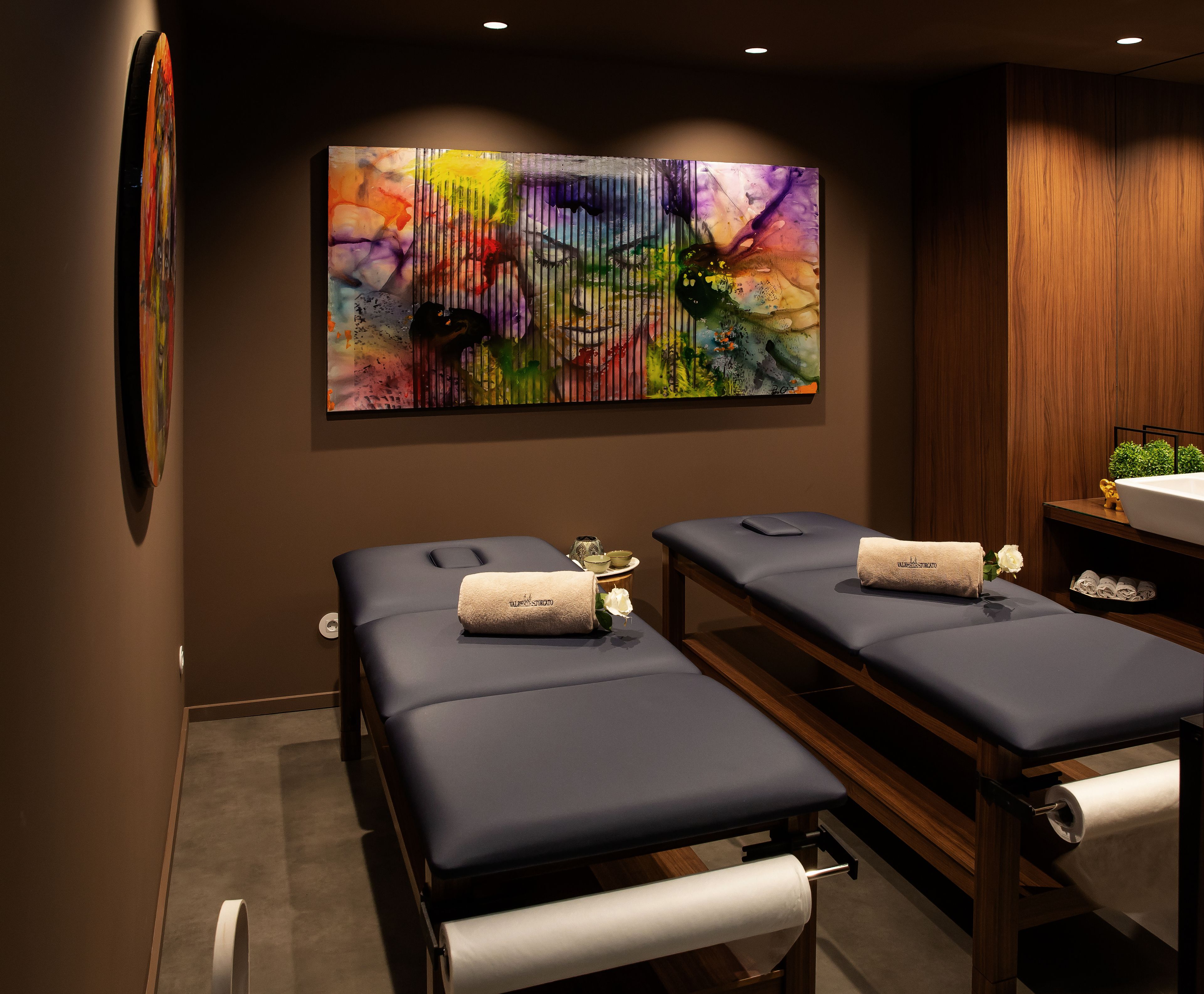 A spa room with two massage tables, a large abstract painting on the wall, and a wooden cabinet with a sink.