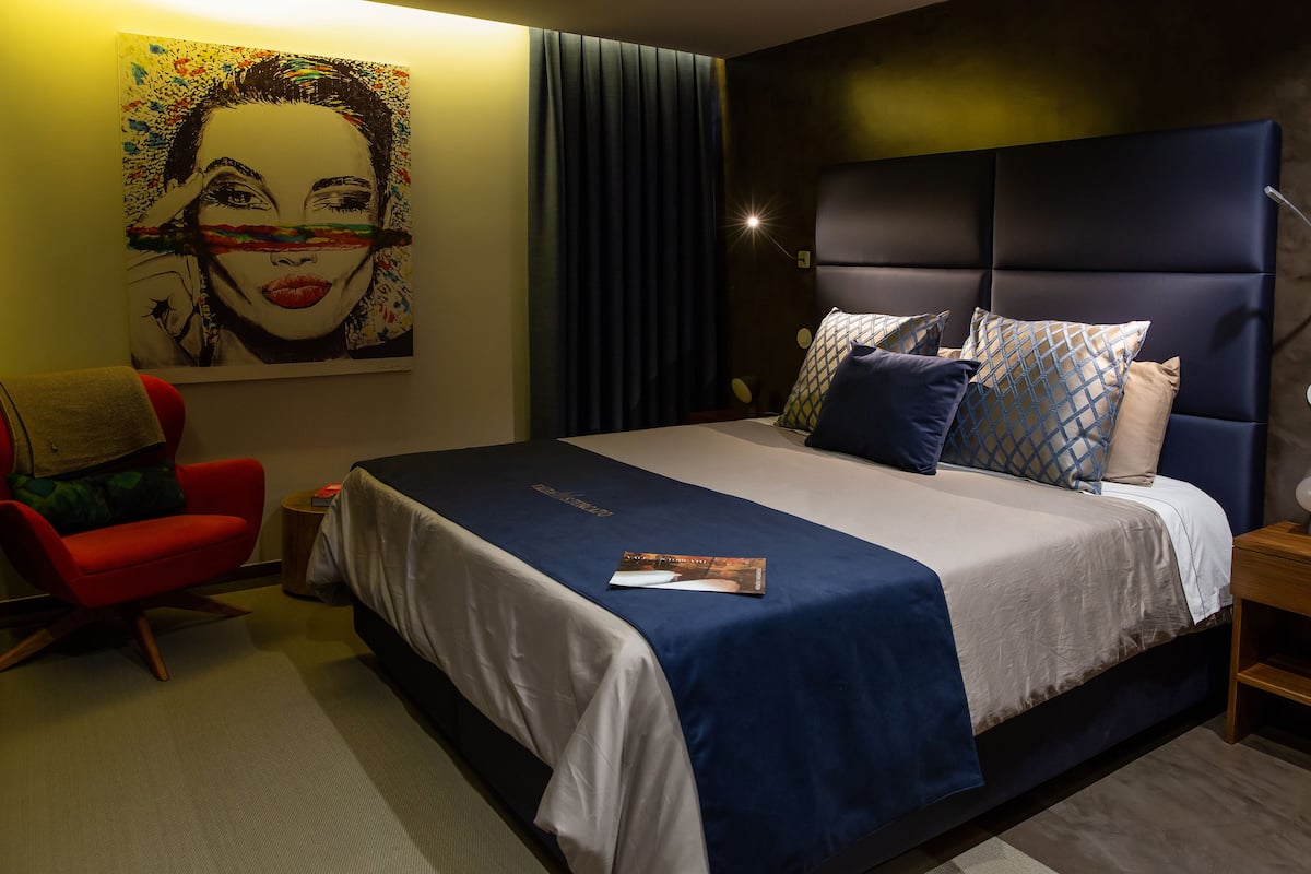 Una habitación de hotel moderna con una cama grande, una silla roja, un cuadro en la pared y una mesita de noche con una lámpara.