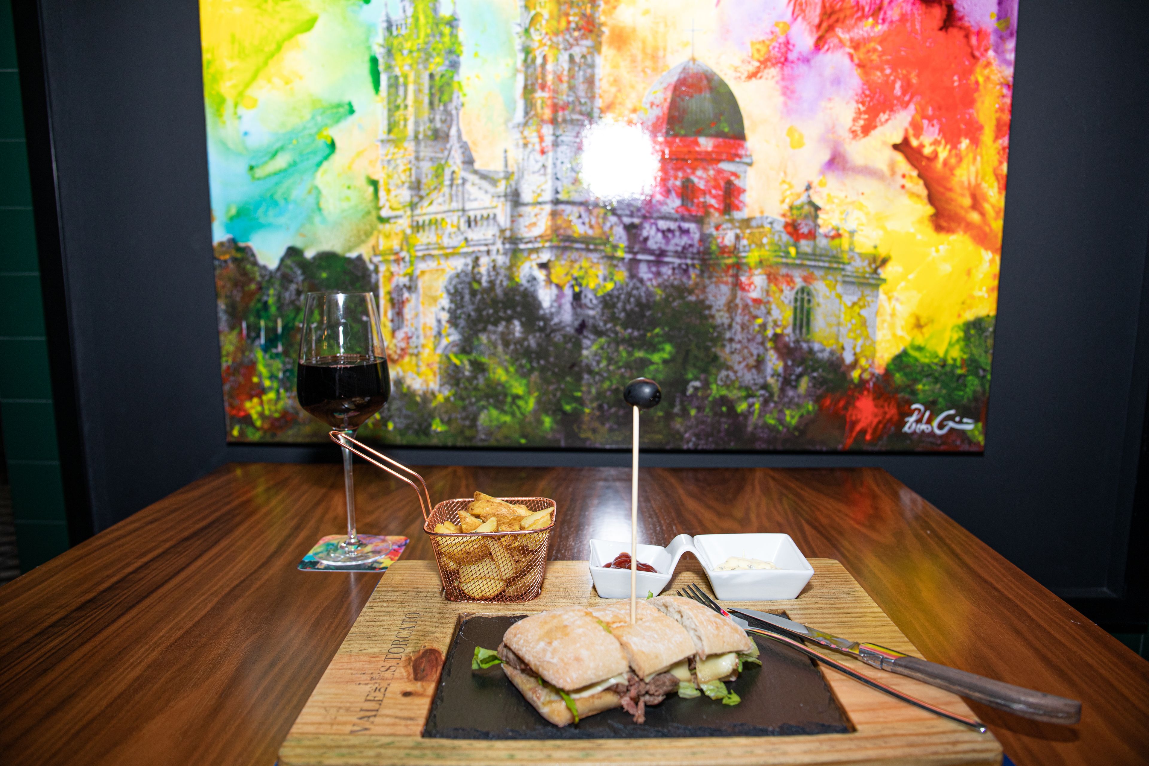 Una mesa de madera con un sándwich, una copa de vino tinto, una cesta de papas fritas y un recipiente pequeño de kétchup.