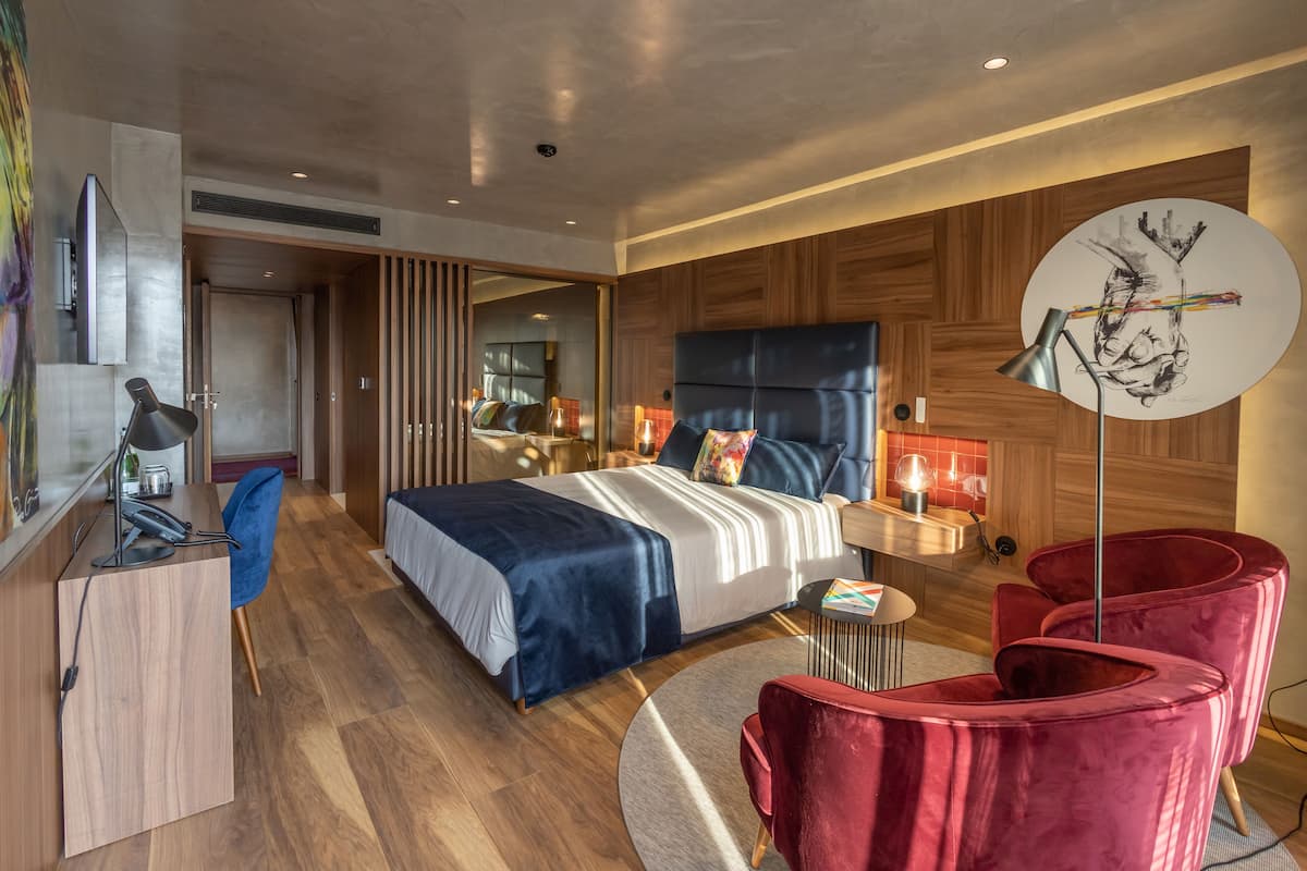 Una habitación de hotel moderna con una cama grande, un escritorio con lámpara, una silla roja y una mesa redonda.