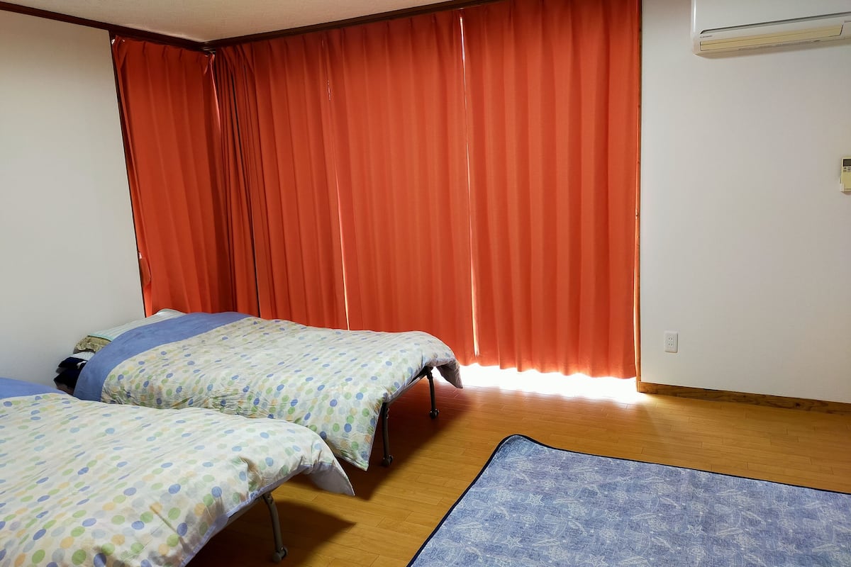 Una habitación con dos camas, un tapete azul estampado, piso de madera, cortinas rojas y un equipo de aire acondicionado.