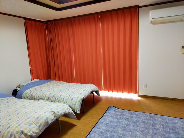 A room with two beds, a blue patterned rug, wooden flooring, red curtains, and an air conditioning unit. - Shirahono Yado Aoiumi (Ishigaki)