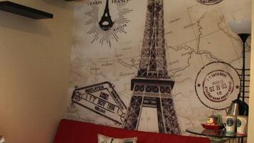 Un dormitorio con un mural temático de mapas que muestra la Torre Eiffel, un sofá rojo y una mesita con una lámpara y adornos.