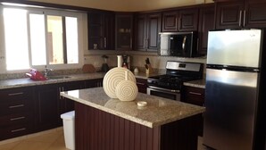 A kitchen with dark wood cabinets, a stainless steel refrigerator, a microwave, and a stove. There is a window with blinds, a sink, and a trash can. - Sosua villa tropical retreat with ocean views (Sosúa)