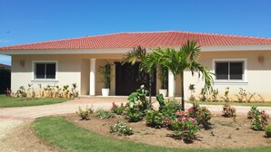 A two-story house with a red-tiled roof, a front porch with a palm tree, and a garden with various plants and flowers. - Sosua villa tropical retreat with ocean views (Sosúa)