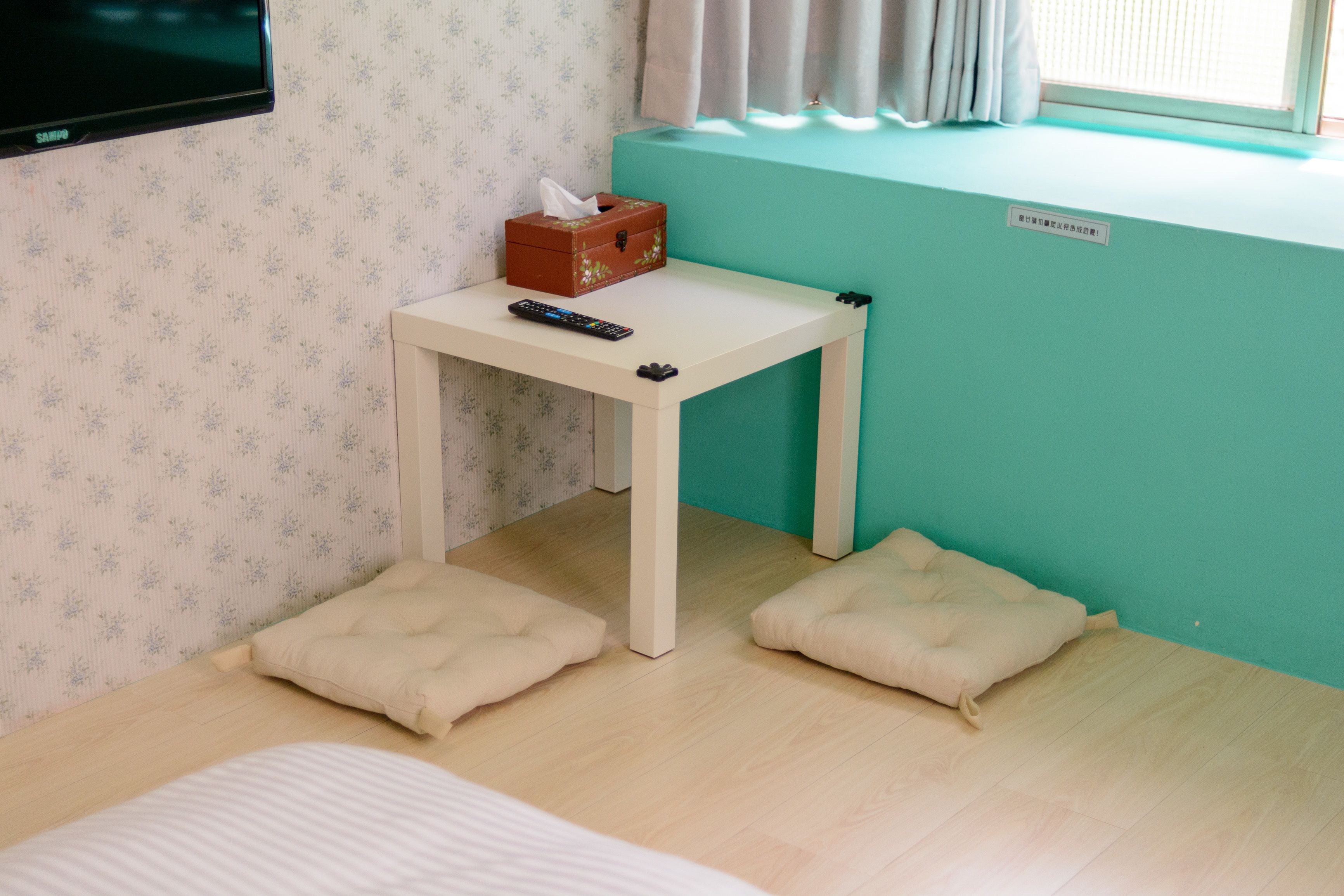 A small room with a bed, a table with a remote control and a tissue box, and a window with curtains.