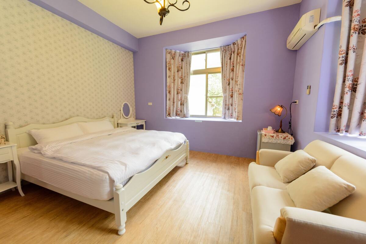 A bedroom with a bed, a nightstand, a lamp, a window with curtains, a sofa, and an air conditioning unit.