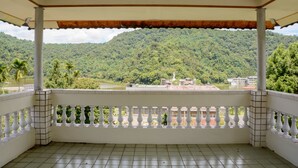 Ein überdachter Balkon mit Fliesenboden und weißer Brüstung, mit Blick auf eine üppige, grüne Hügelkuppe und ein Gewässer.