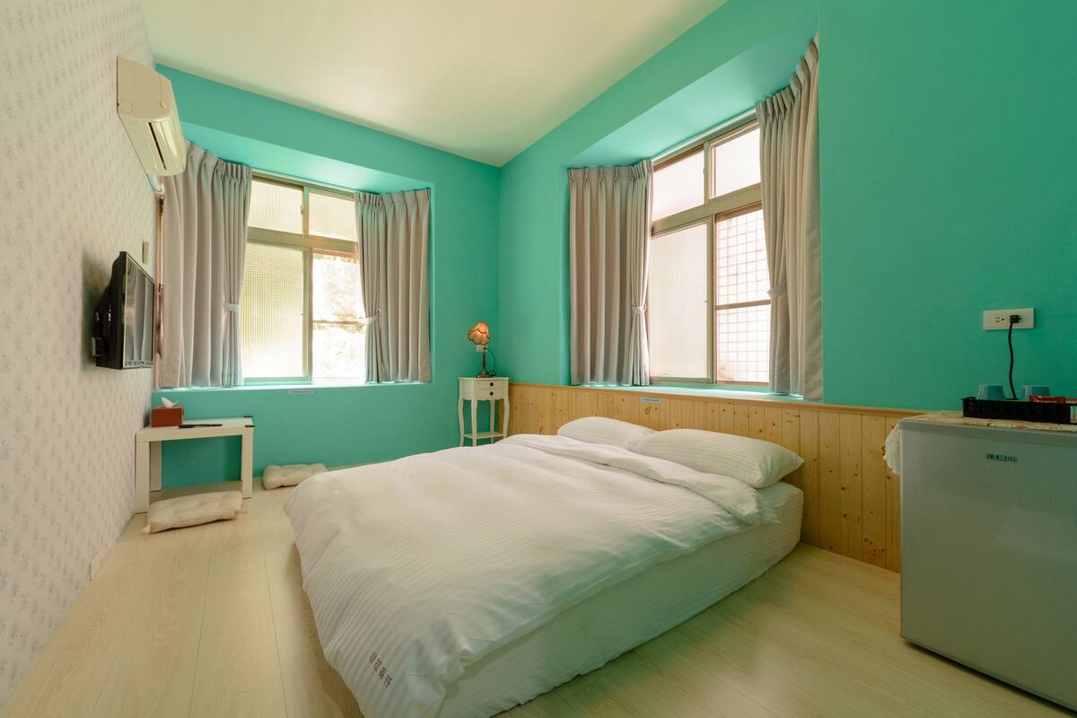 A room with teal walls, a bed, a small table, a lamp, and a refrigerator.