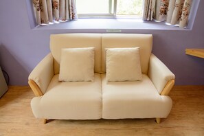 A beige sofa with two cushions in a room with wooden flooring, a window with floral curtains, and a purple wall. - Bella Forest (Puli)