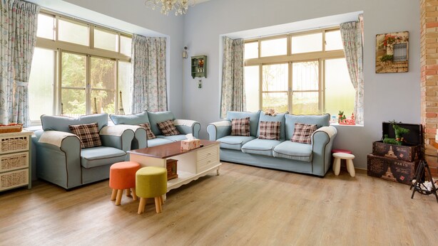 Ein geräumiges Wohnzimmer mit zwei hellblauen Sofas, einem Holztisch und einem kleinen Hocker. Große Fenster mit floralem Vorhangstoff, eine Kristallleuchter und eine Ziegelwand mit Dekorationselementen.