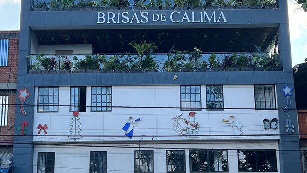 Een hotel genaamd 'BRISAS DE CALIMA' met een modern design, met balkons met planten, een etalage met de teksten 'Pina colada' en 'naturais plus', en een heldere blauwe lucht op de achtergrond.