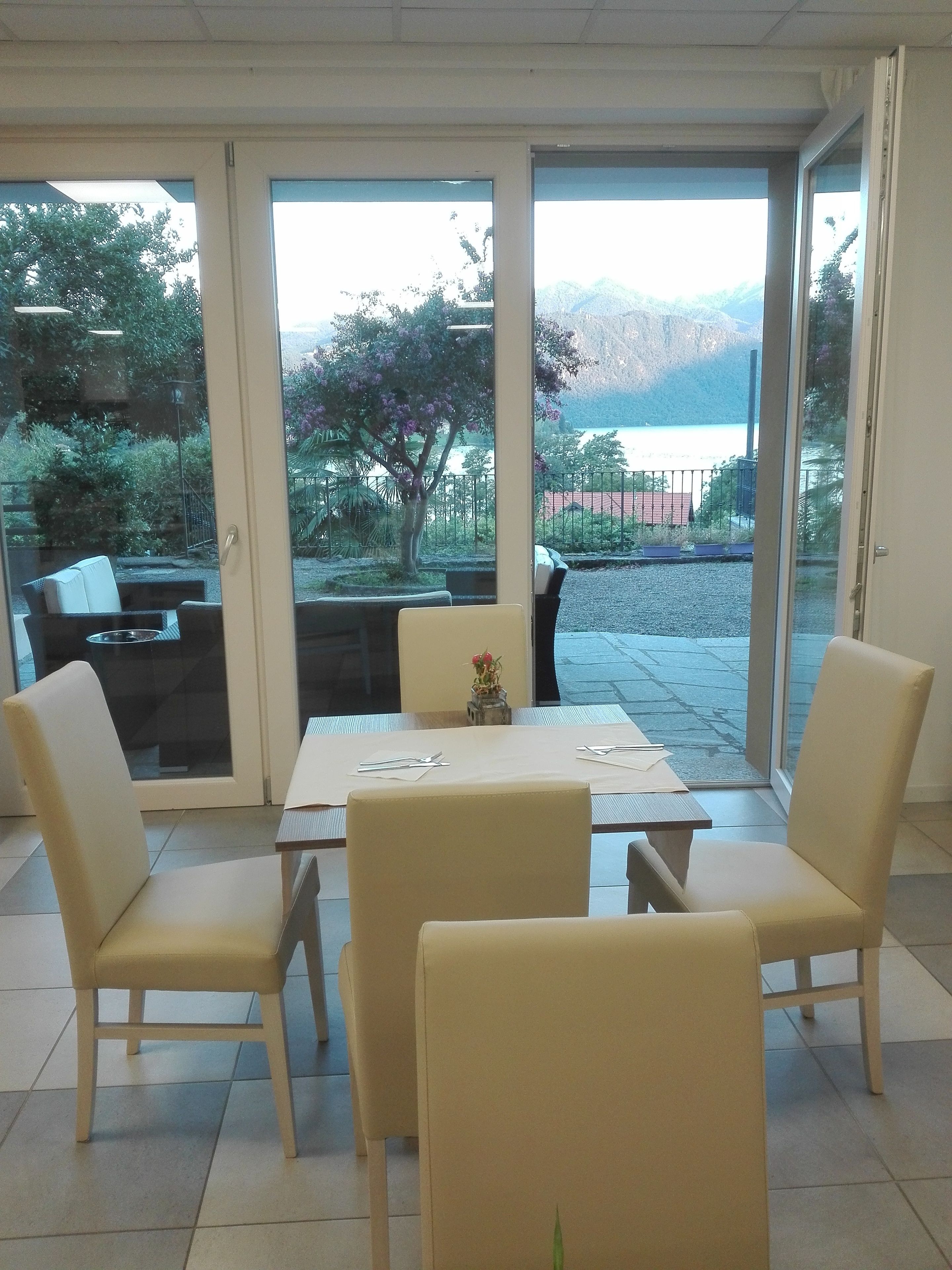A dining area with a table and four chairs, a small vase with flowers, and a view of a lake and mountains through large glass doors.