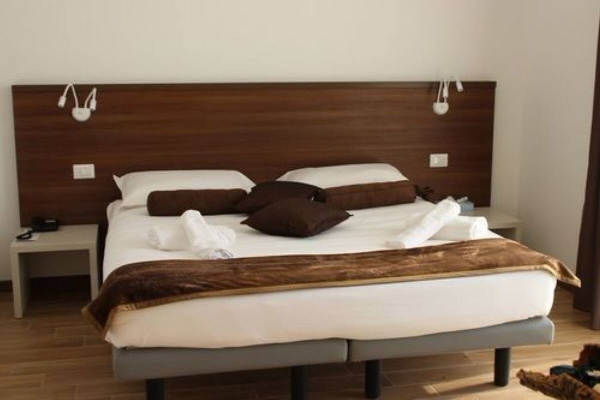 Un lit bien fait, avec une tête de lit marron, du linge de lit blanc et un plaid marron. On y trouve deux tables de chevet avec des lampes, ainsi qu’une petite table de nuit avec un téléphone.