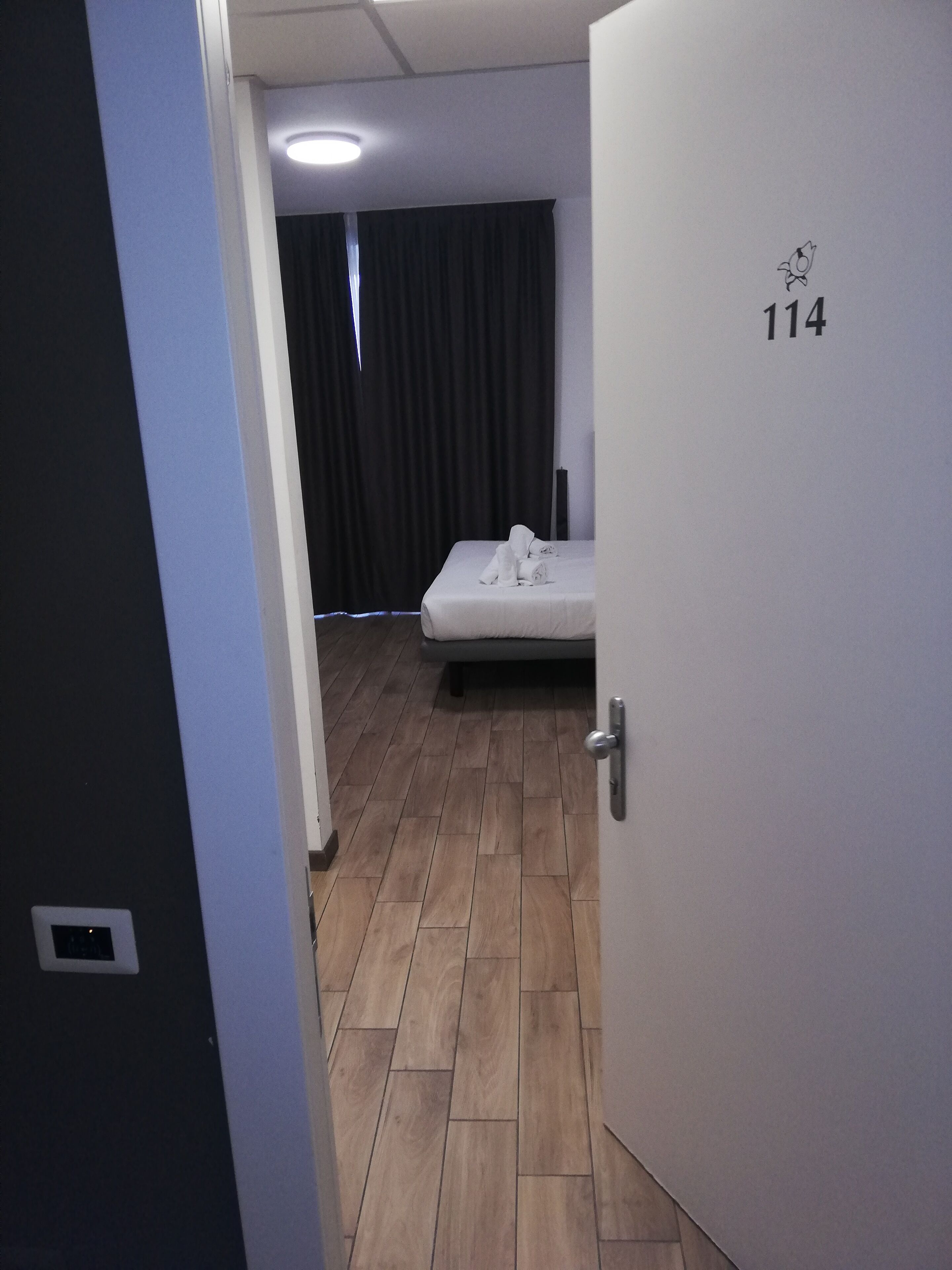 Habitación de hotel con una cama, una puerta con el número 114, una ventana con cortinas y suelo de madera.