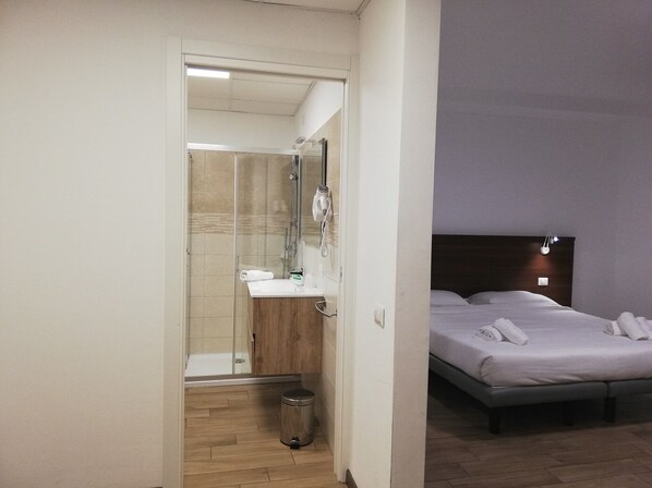 Una habitación de hotel con ducha, tocador de madera, cama con ropa blanca y mesita de noche.
