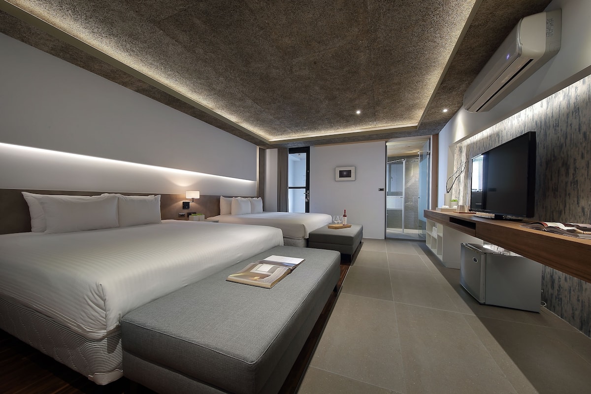 A modern hotel room with a large bed, a smaller sofa, a desk with a TV, and a kitchenette.