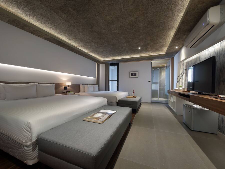 A modern hotel room with a large bed, a smaller sofa, a desk with a TV, and a kitchenette.
