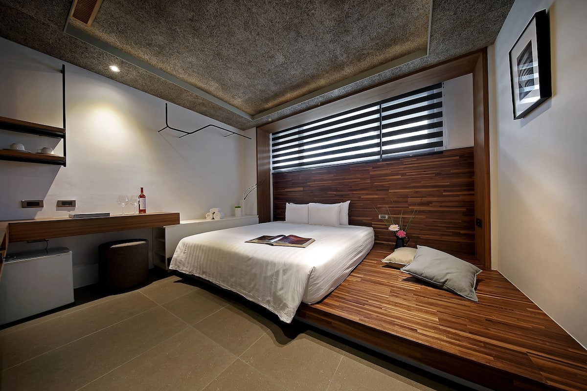 A modern bedroom with a wooden headboard, a bed with white bedding, a bedside table with a lamp, and a small bedside plant.