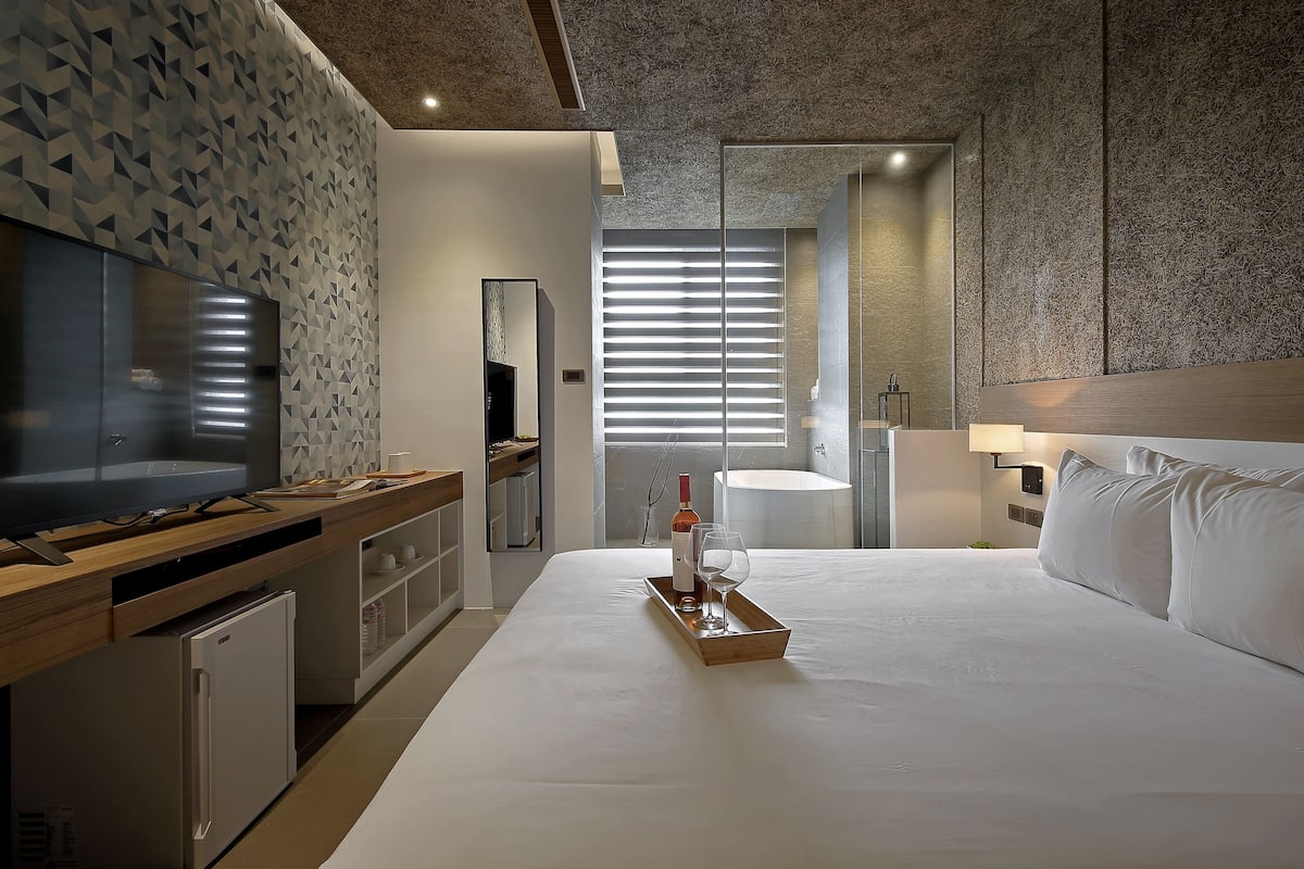 A modern hotel room with a large bed, a wooden desk with a TV, a mini-fridge, and a tray with a wine bottle and glass.