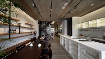 A modern restaurant with a long wooden table, seating, and a kitchen area with shelves of dishes and glasses.