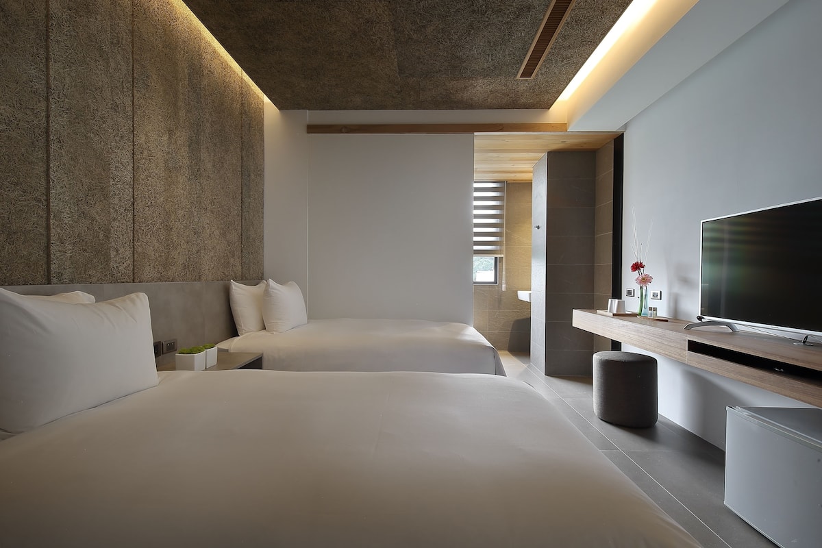 A modern hotel room with two beds, a flat-screen TV, a small table, and a minimalist design.