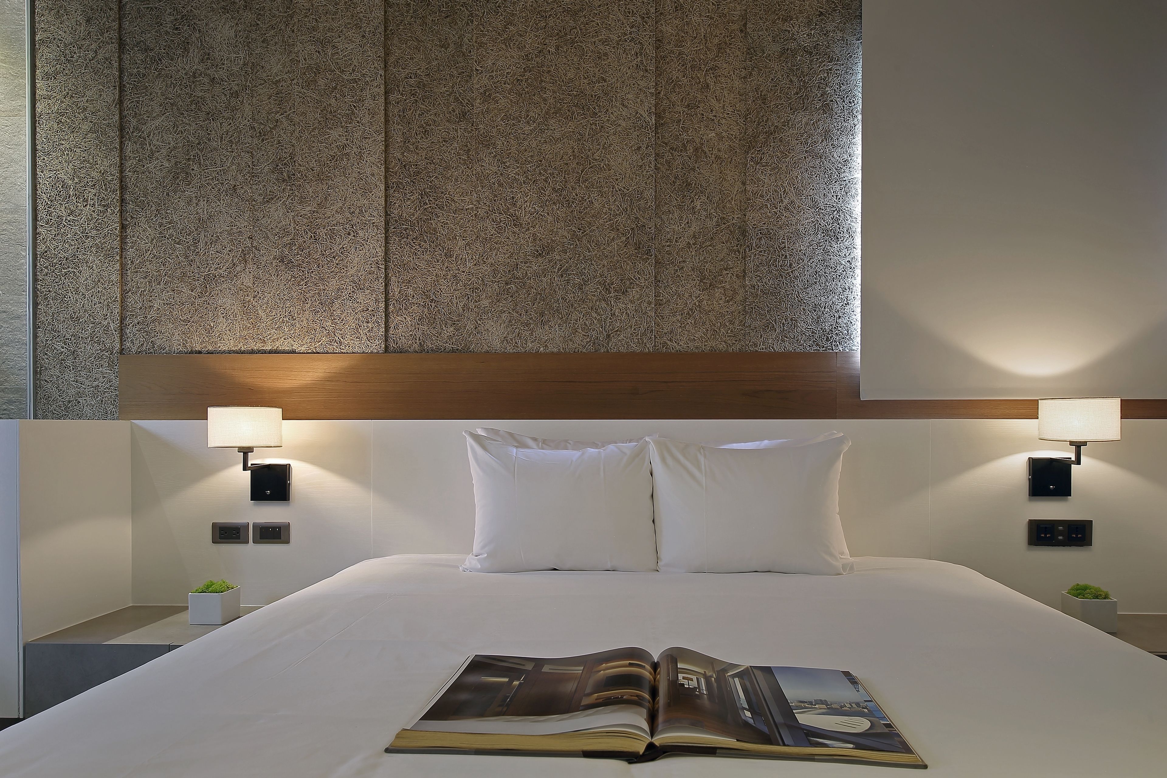 A neatly made bed with white linens and pillows, a magazine on the bed, and two bedside lamps.