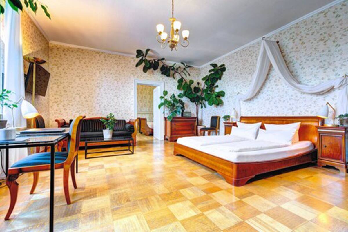 A hotel room with a wooden floor, a bed with white linens, a desk with a chair, a small table, and a potted plant.