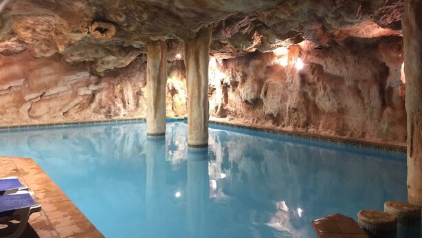 An indoor pool with a rocky cave-like ceiling, clear blue water, and stone pillars. - Aparthotel Sole Bello (Torrevieja)