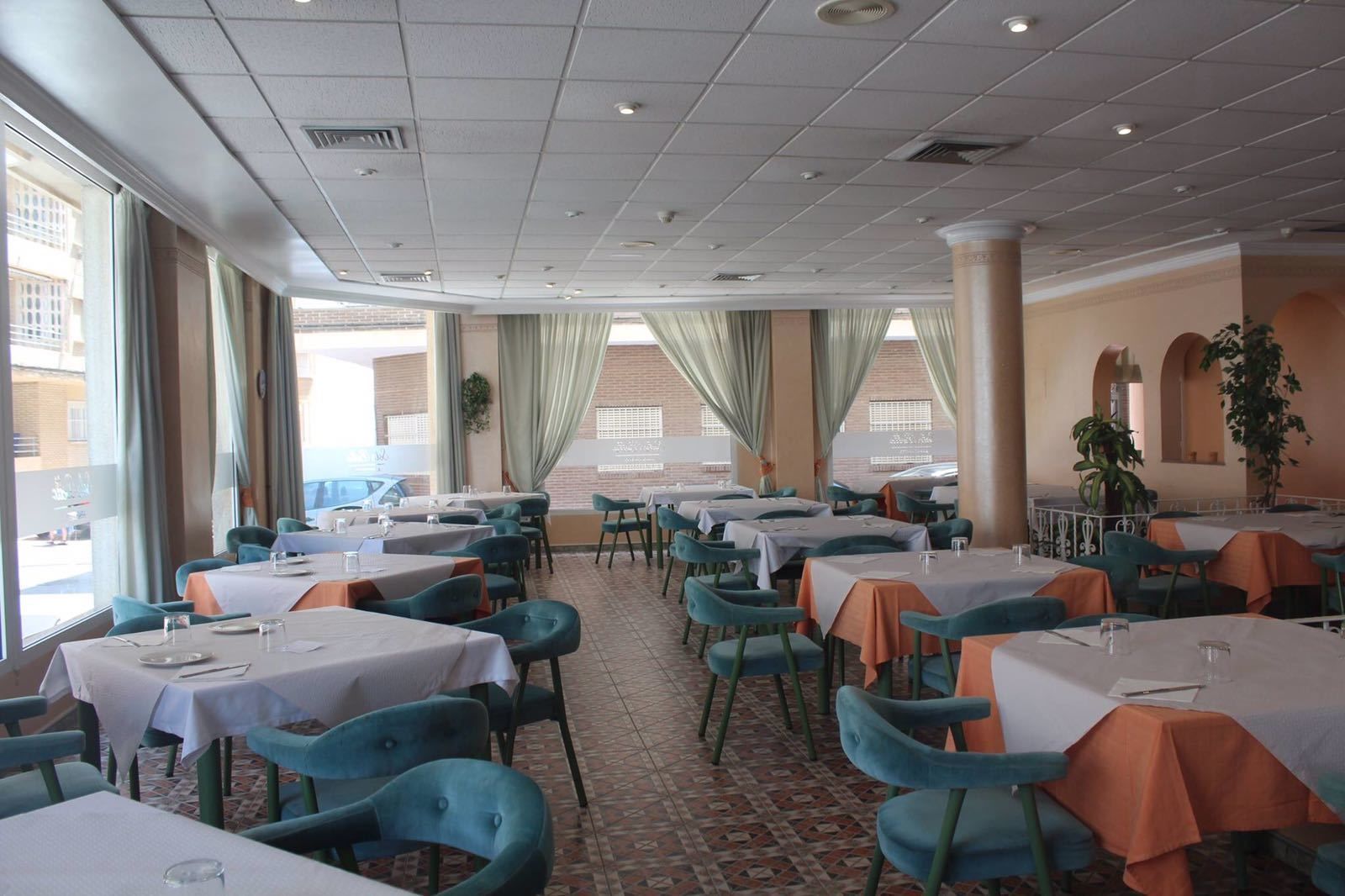 A restaurant with multiple tables and chairs, large windows with curtains, and a tiled floor.