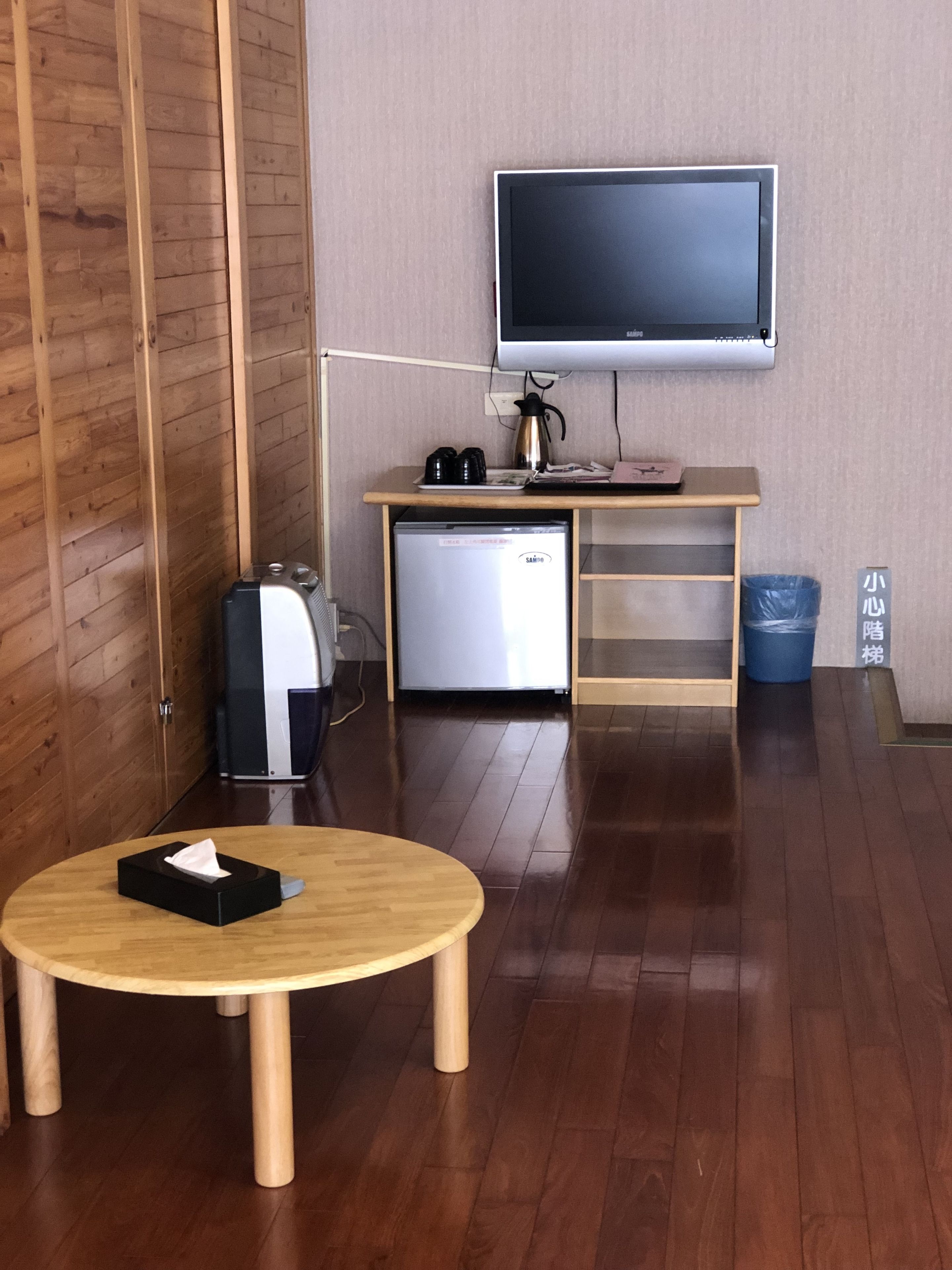 Una habitación compacta con piso de madera, una pequeña kitchenette con refrigerador, un televisor montado en la pared, una mesa de madera con caja de pañuelos y un cesto de basura.