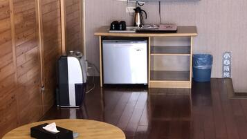 Una habitación compacta con piso de madera, una pequeña kitchenette con refrigerador, un televisor montado en la pared, una mesa de madera con caja de pañuelos y un cesto de basura.