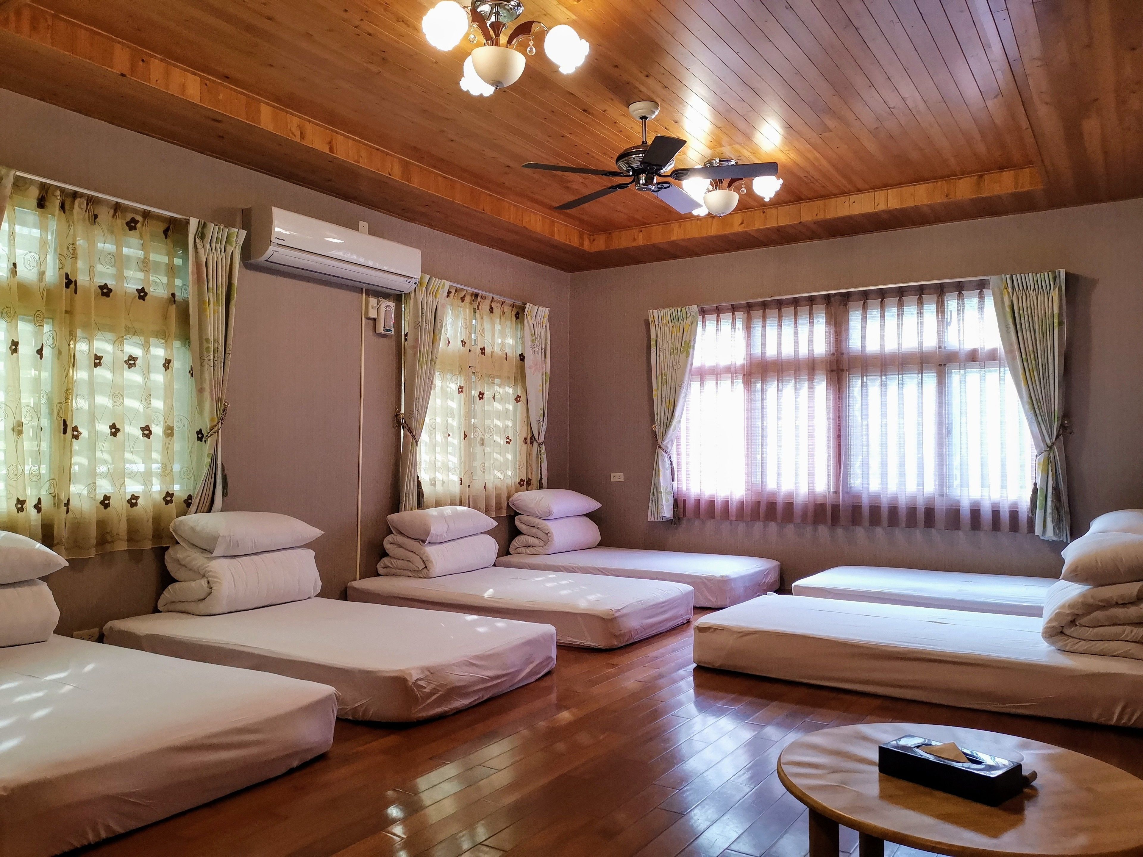 A room with multiple beds, wooden flooring, ceiling fans, and large windows with curtains.