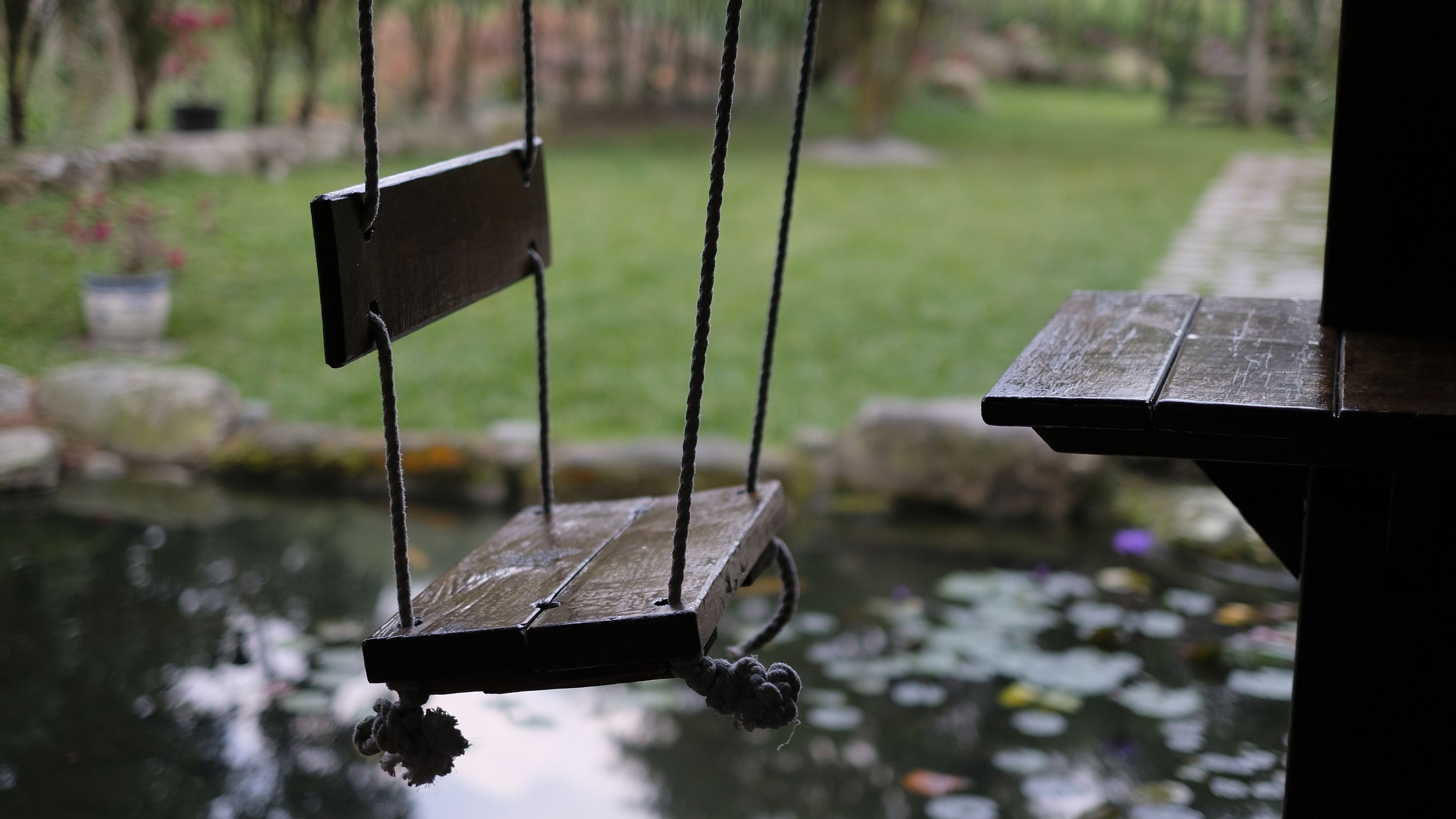 Una hamaca de madera suspendida sobre un estanque, con un sendero de piedra al fondo.