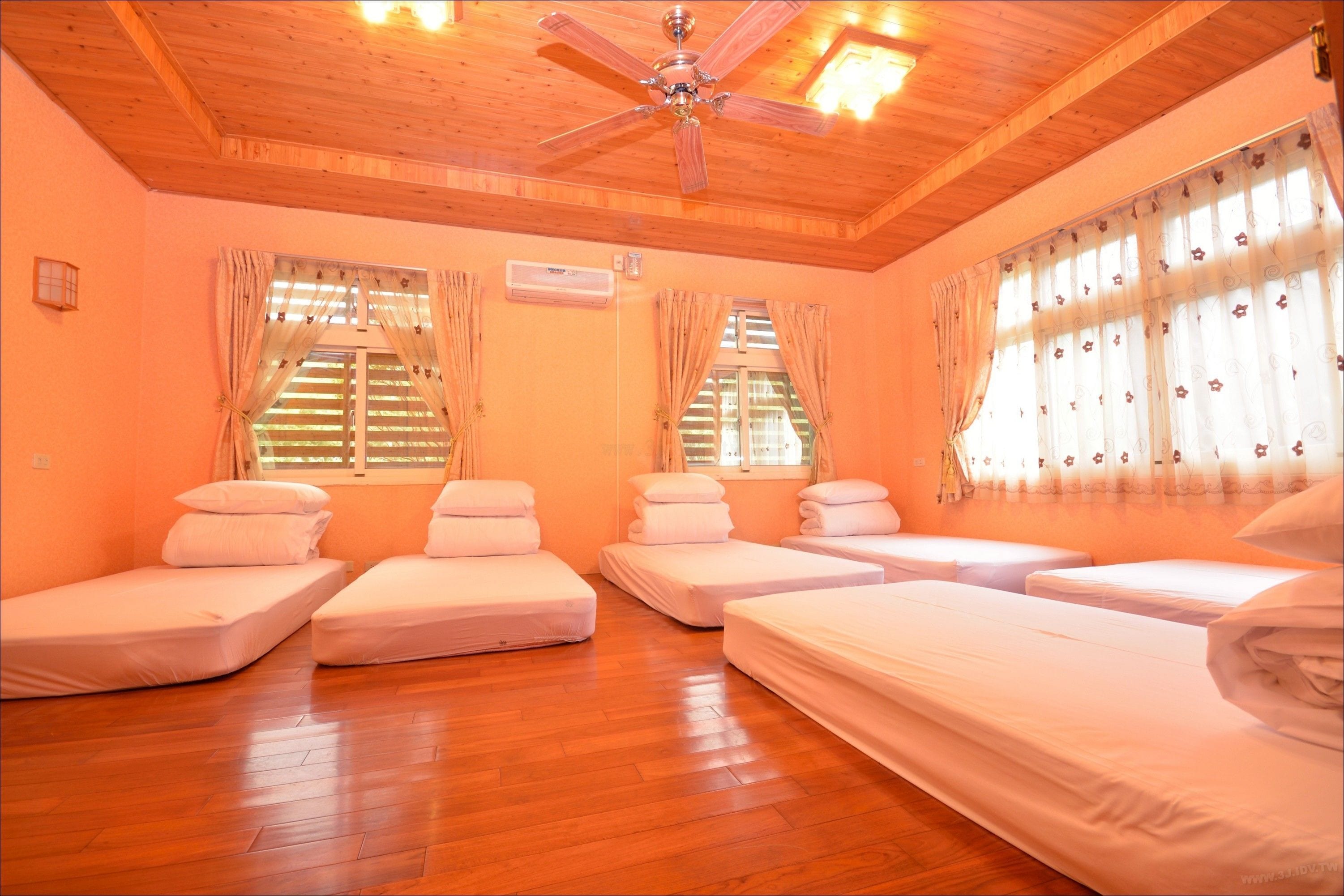 Habitación de hotel con cuatro camas individuales, suelo de madera, ventilador de techo y ventanas con cortinas.