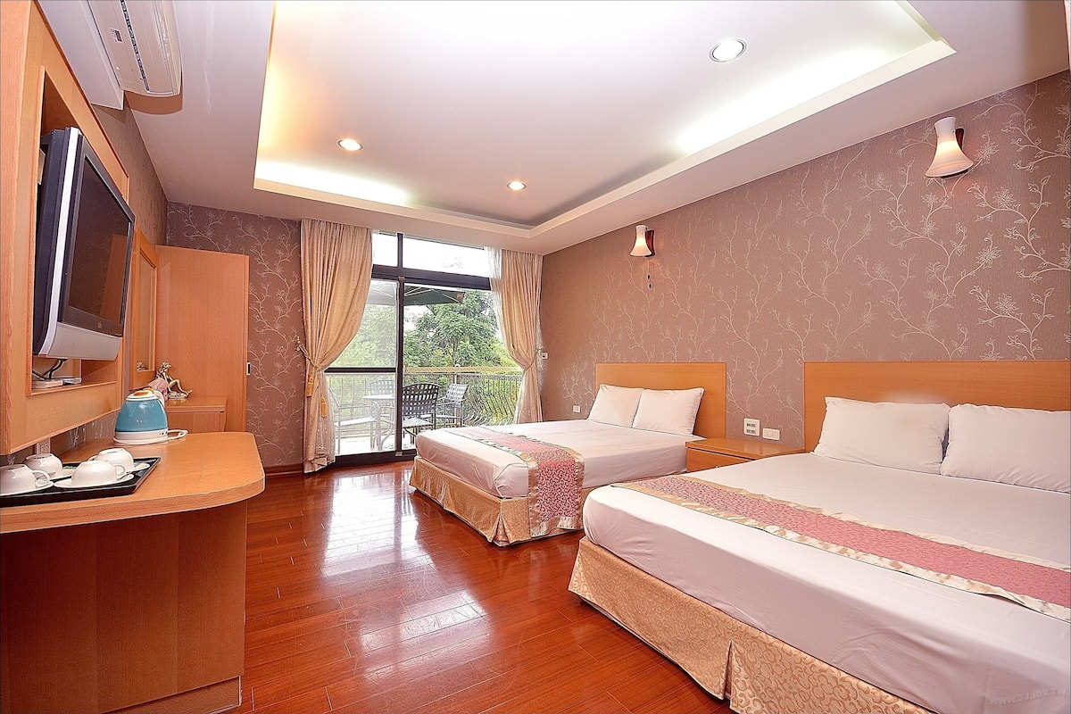 A hotel room with two beds, a desk with a kettle, and a large window.