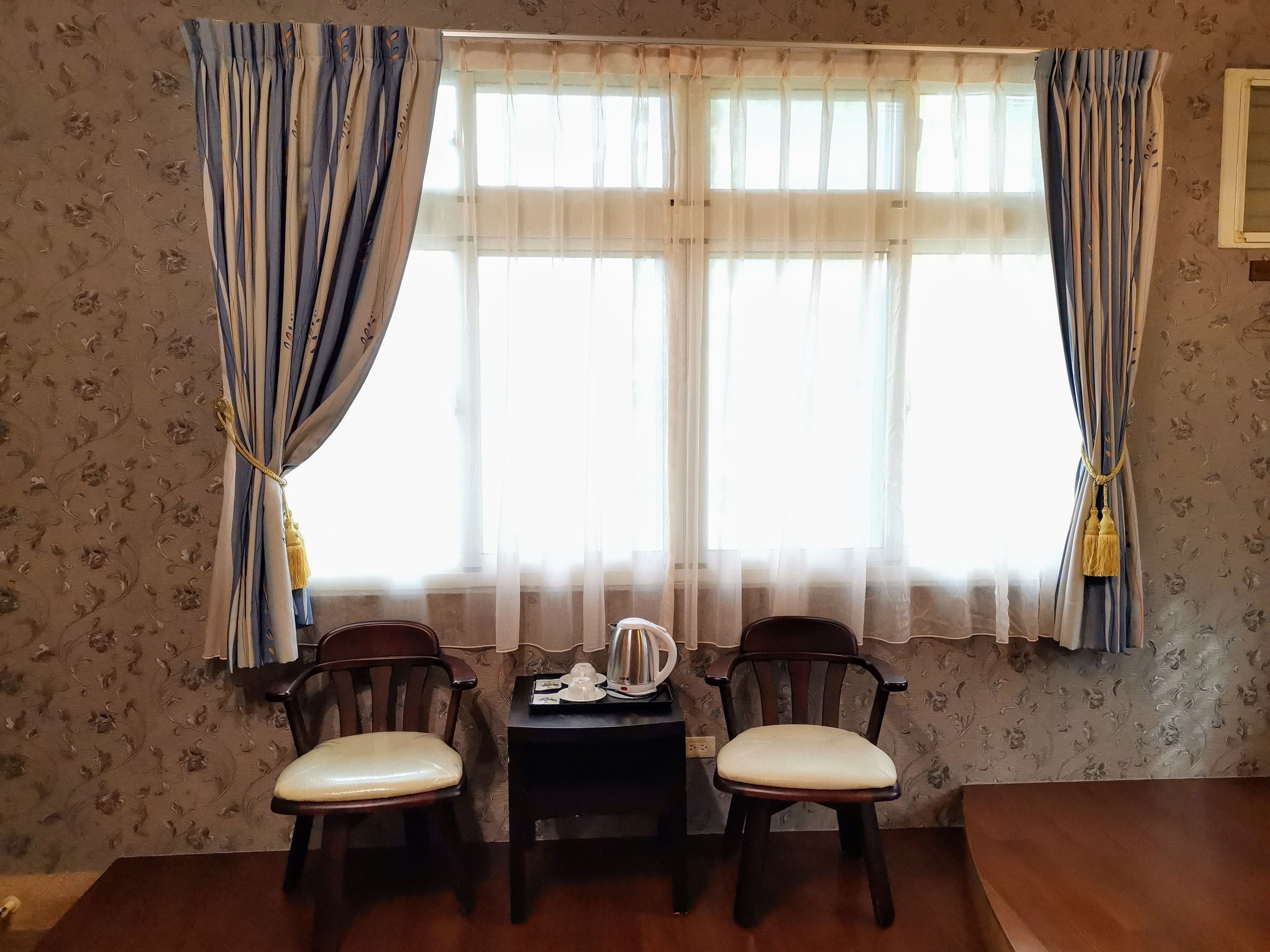 A room with two wooden chairs, a small table with a kettle, and a window with curtains.